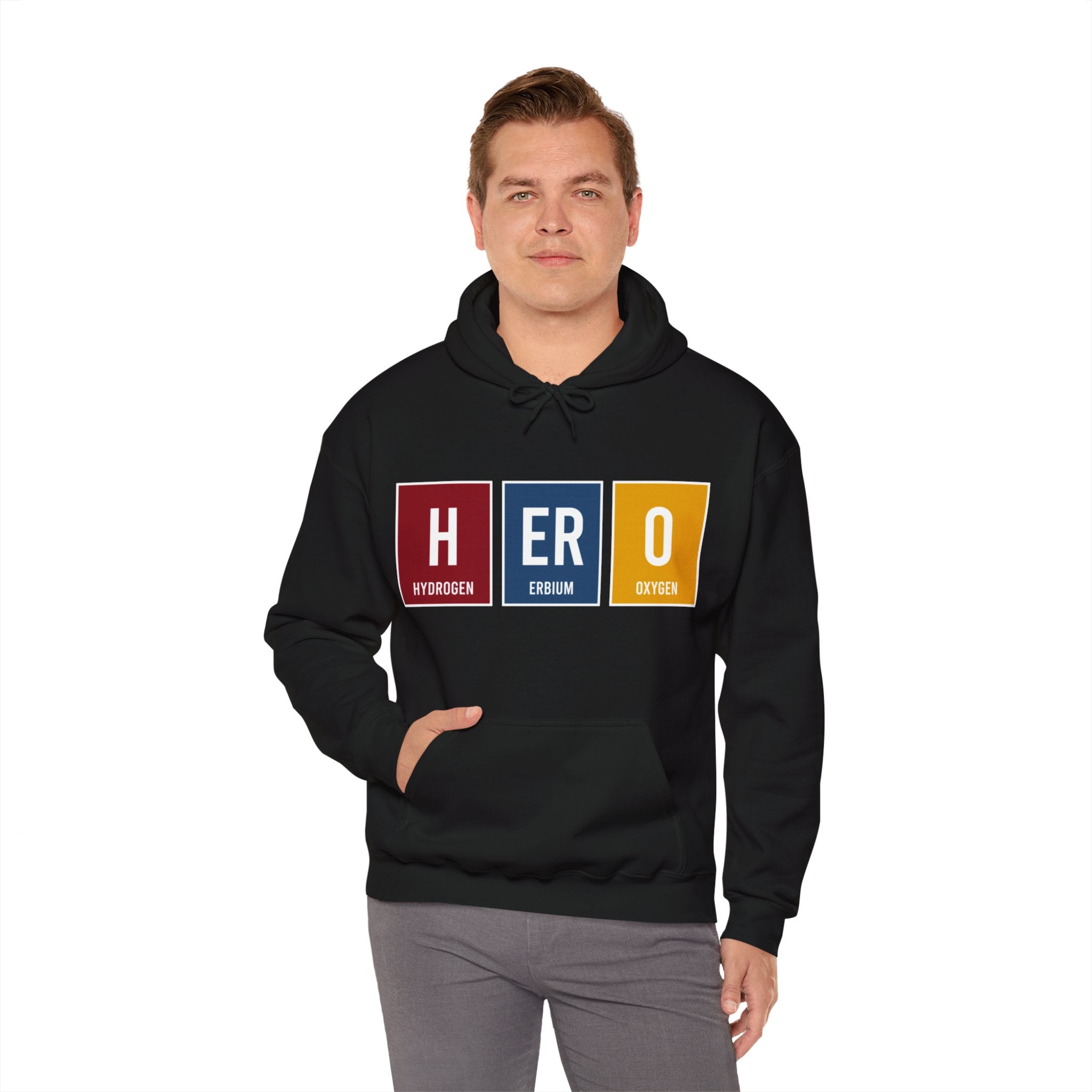 A person in a HERO - Hooded Sweatshirt stands with hands in the front pocket. The hoodie displays the text "HERO" using periodic table elements: Hydrogen, Erbium, and Oxygen, showcasing HERO designs that epitomize heroism.