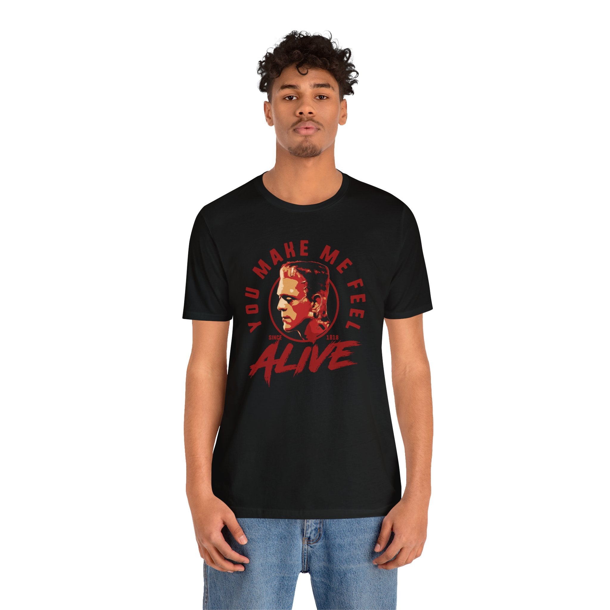 A young man with curly hair wearing a black unisex tee with a red and white "You Make Me Feel Alive" print design and text, paired with blue jeans, standing against a white background.