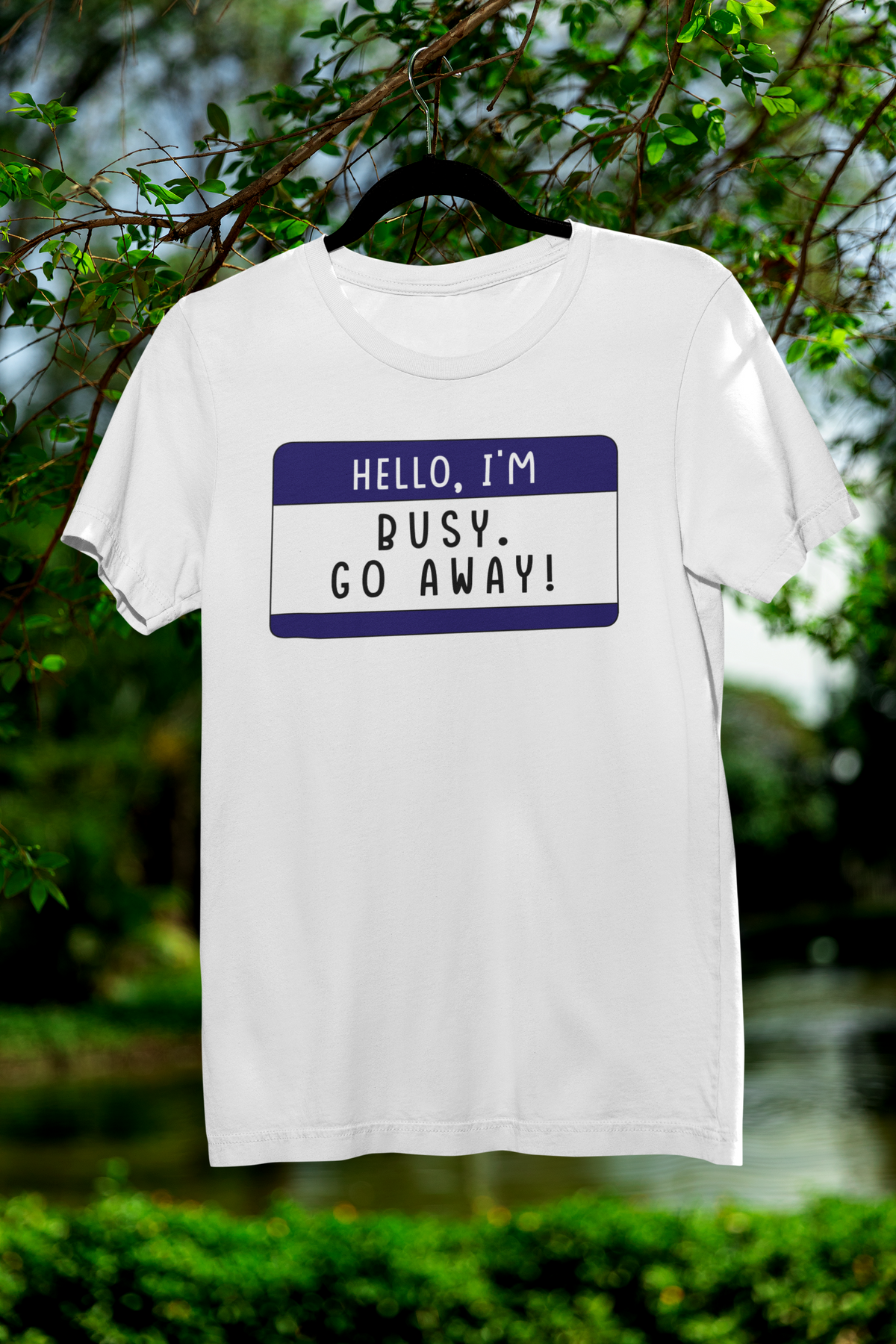 Hello I'm Busy Go Away name tag T-shirt in white, hanging outdoors among greenery, sarcastic gift tee