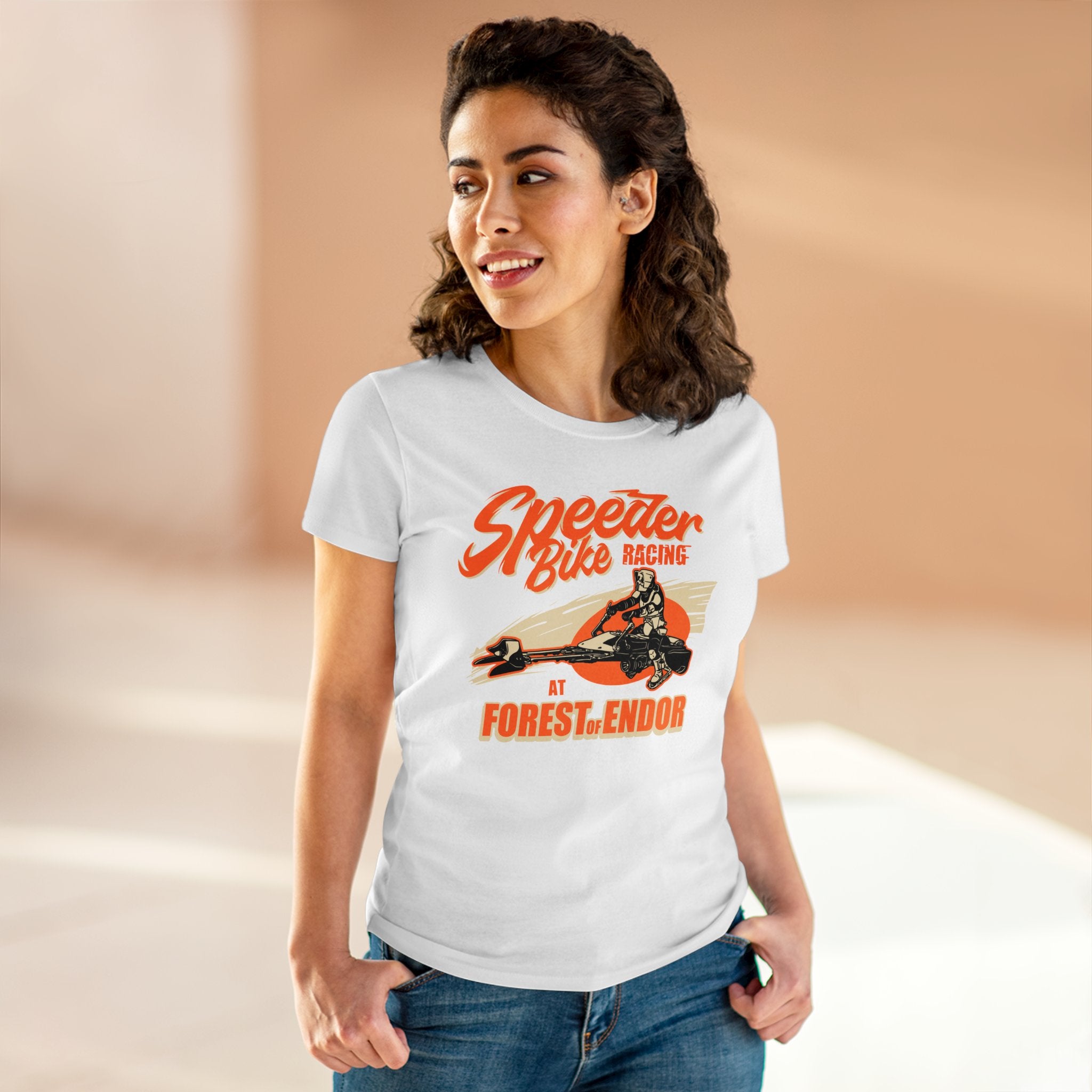 A woman with curly hair is wearing a white, pre-shrunk cotton Speeder Bike Racing - Women's Tee with an orange and black graphic. She stands smiling in a light-filled room.