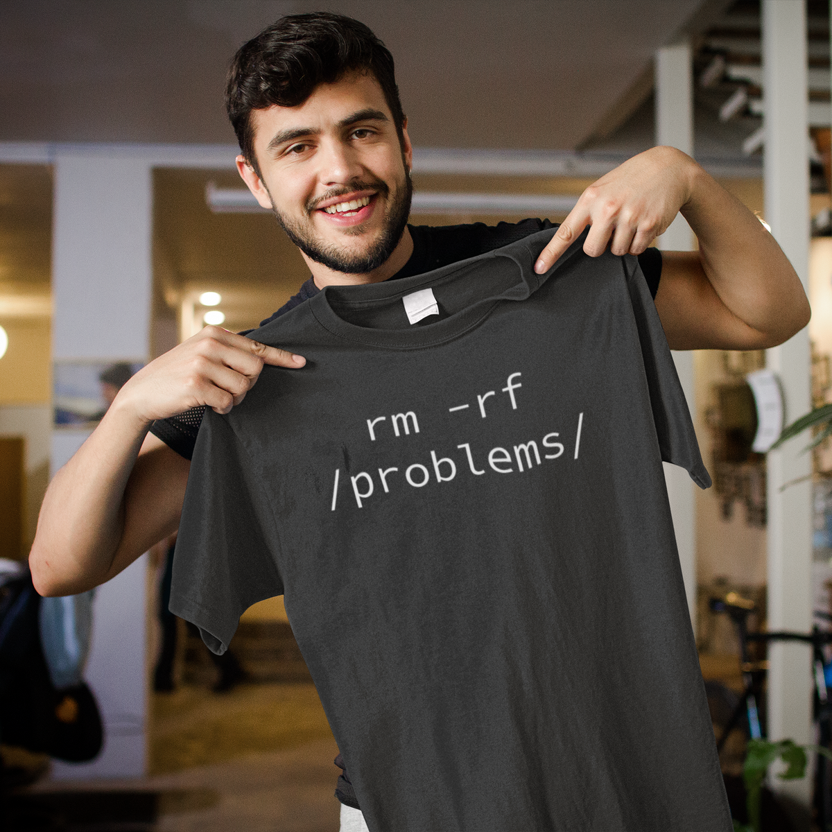 'rm -rf /problems/' t-shirt in black, man holding it up indoors, funny gift for programmers