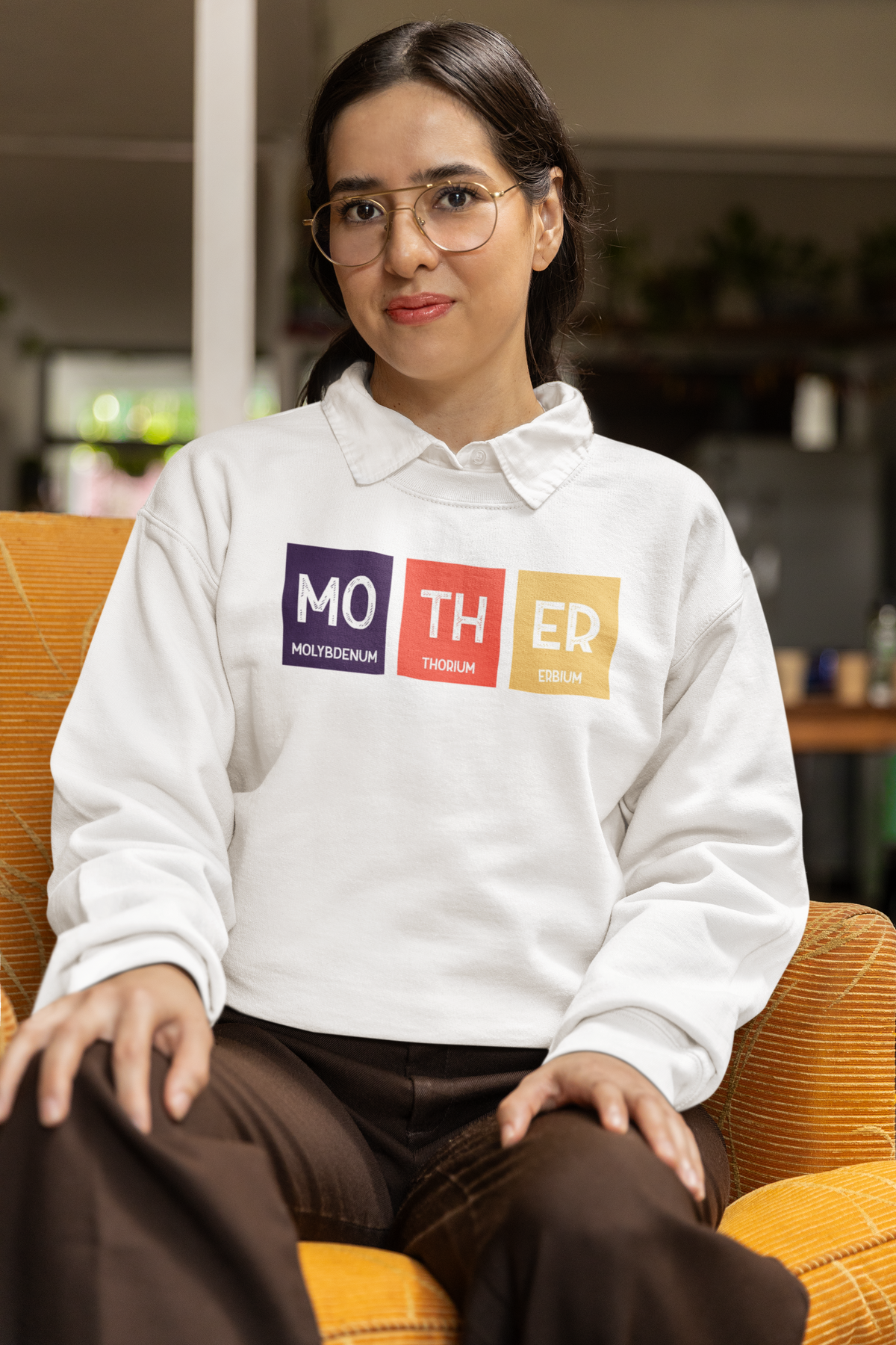 'Mo-TH-ER' periodic table sweatshirt in white, woman sitting in an armchair wearing it, cozy photo