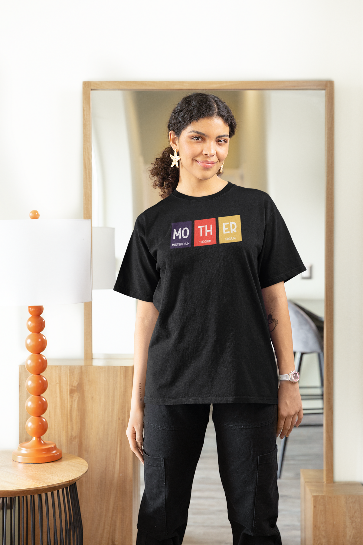 'Mother' periodic table women's tee in black, woman standing at home, casual lifestyle photo