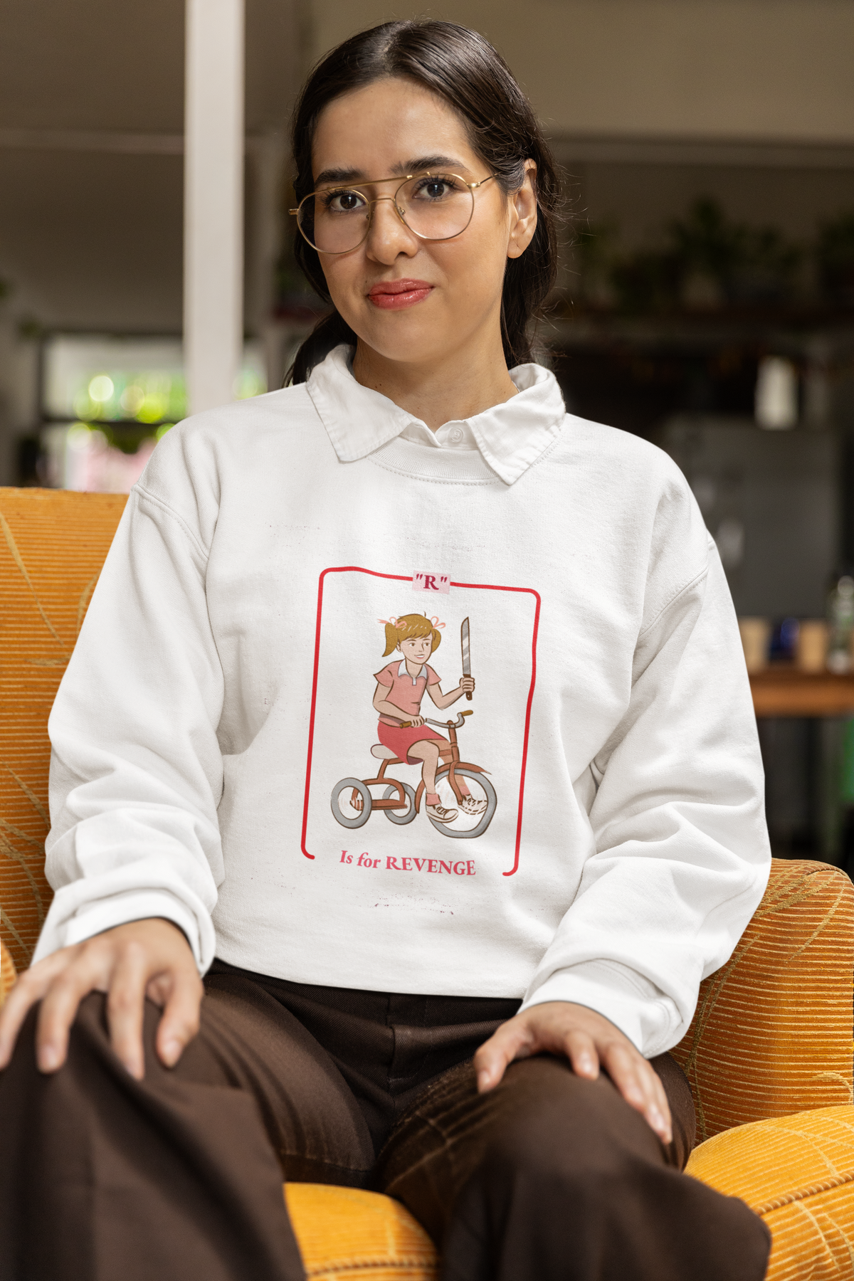 'R is for Revenge' tricycle girl graphic sweatshirt in white, woman wearing it seated indoors, funny gift