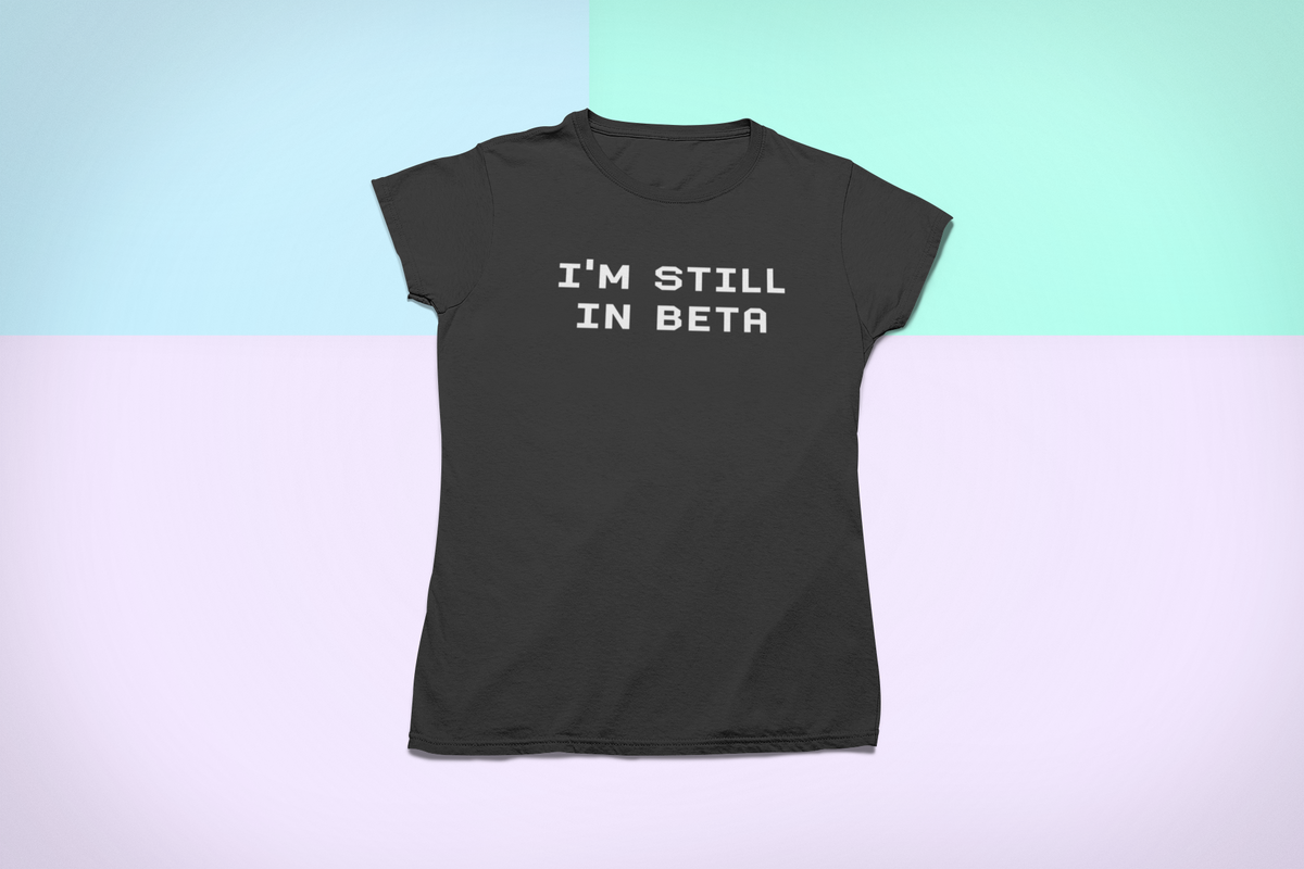 "I'm Still In Beta" typography t-shirt in black, flat lay on pastel background, funny developer pun gift