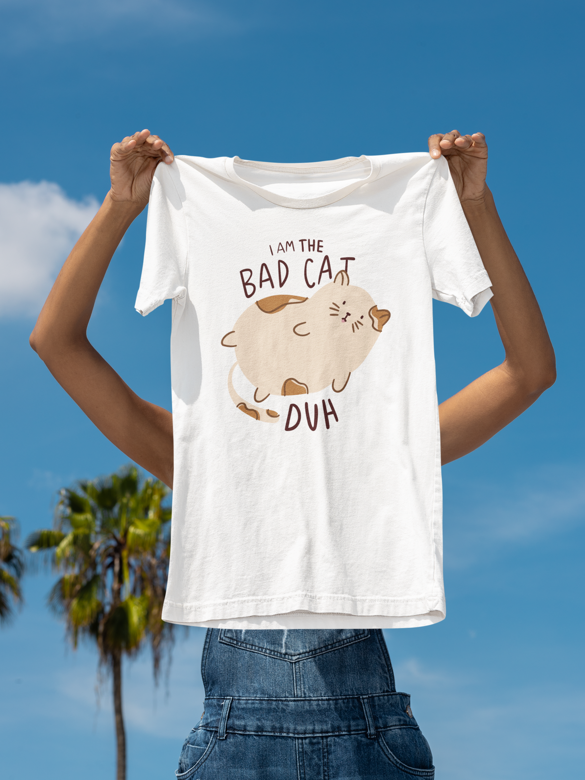 'I Am the Bad Cat Duh' T-shirt in white, woman holding it up at the beach, funny cat lover gift
