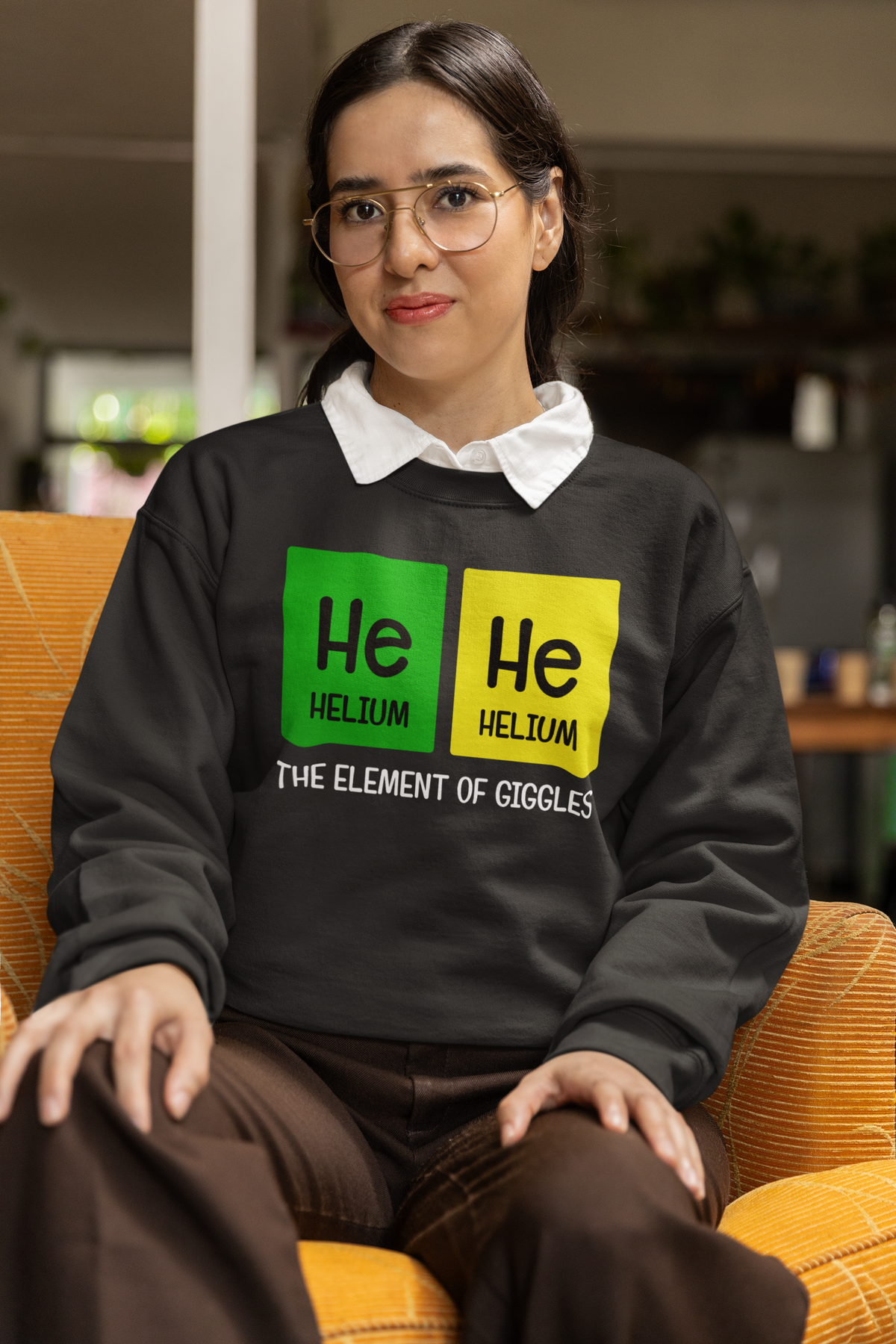 'He-He: The Element of Giggles' sweatshirt in black, woman sitting indoors, funny chemistry gift