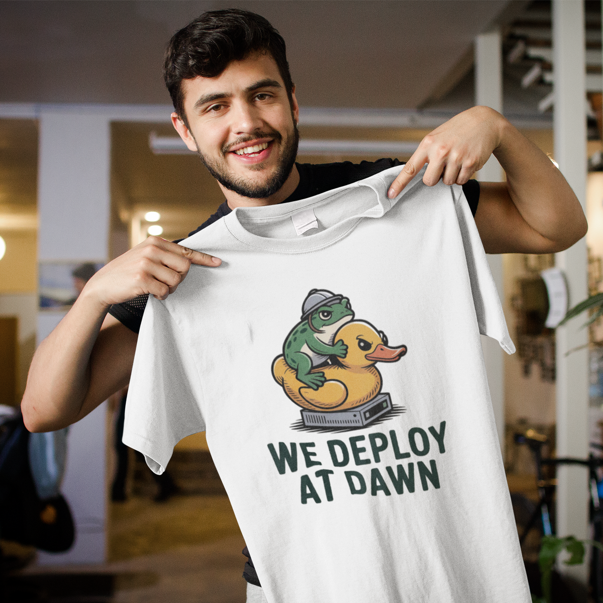 'We Deploy at Dawn' tee in white, man holding it up indoors, funny gift for developers