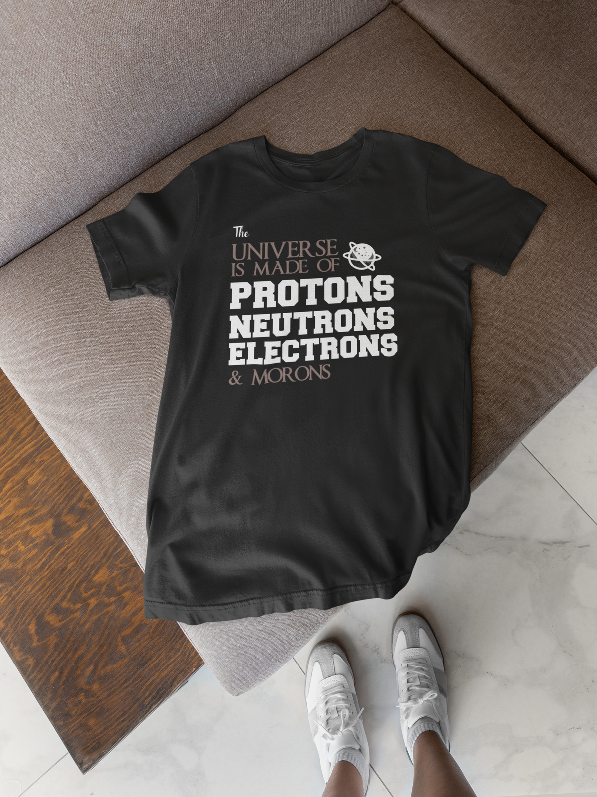 'The Universe Is Made Of...Morons' t-shirt in black, flat lay on a wooden floor, funny sarcastic gift shirt