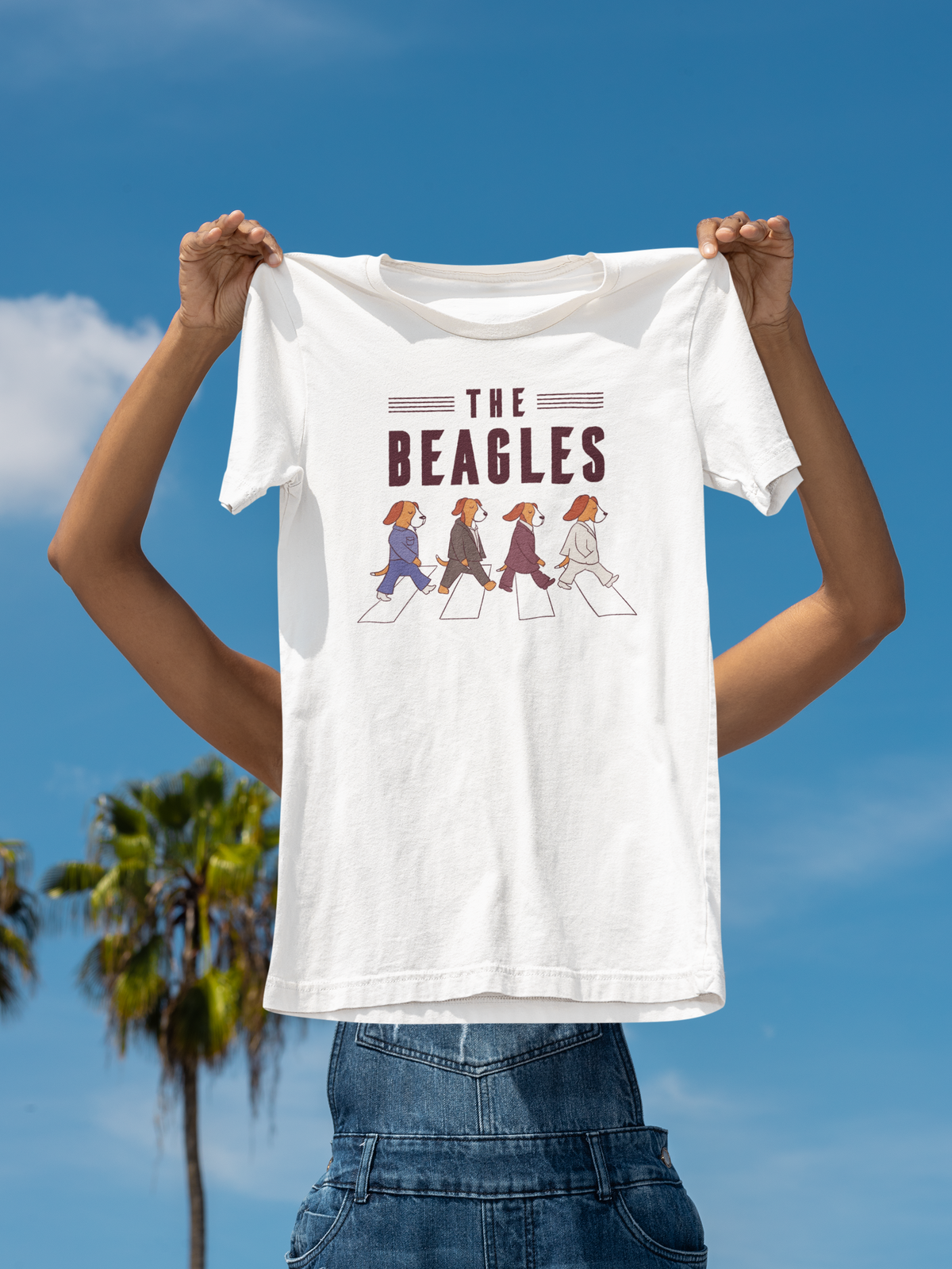 'The Beagles' t-shirt in white, woman holding it up against a palm tree sky backdrop