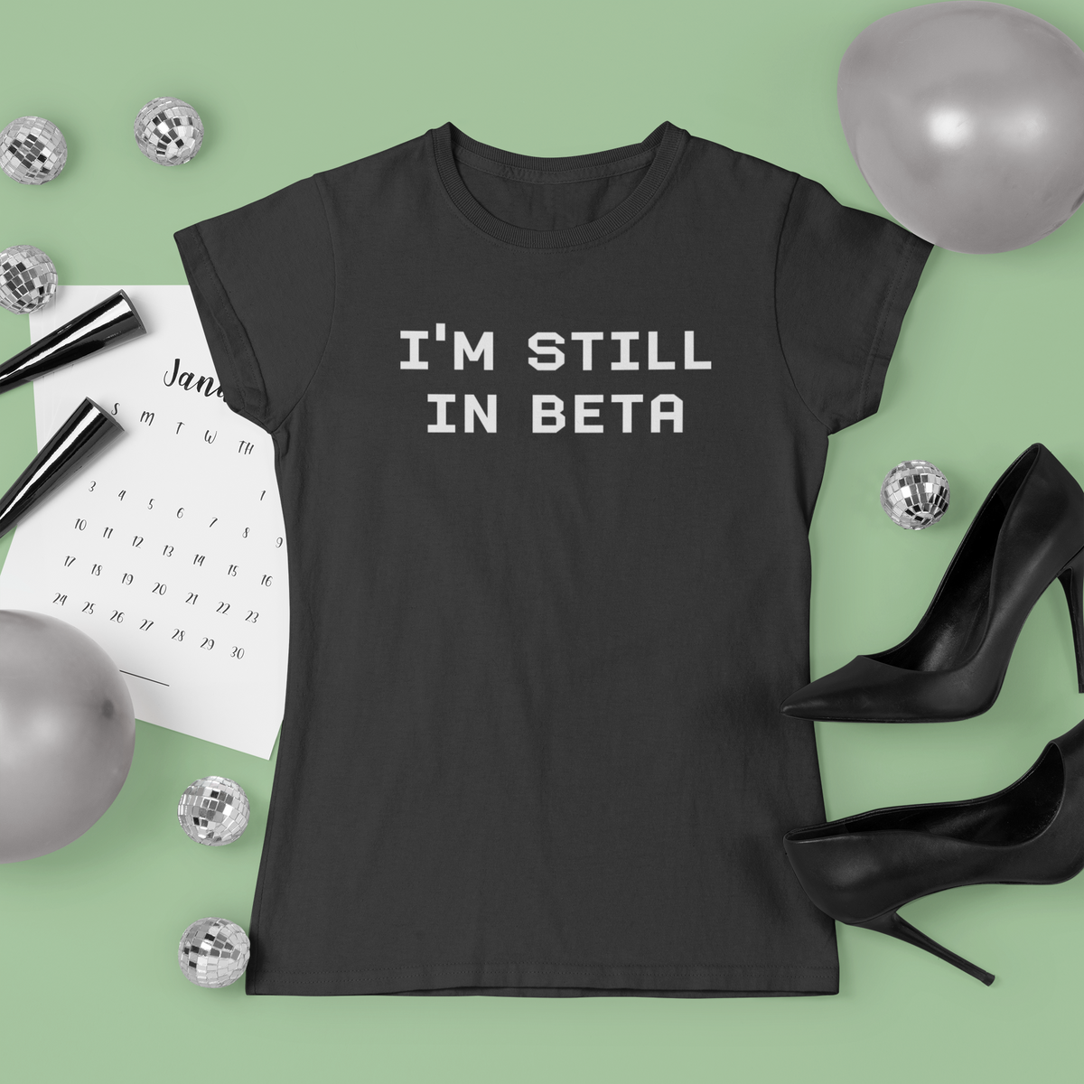 "I'm Still In Beta" typography t-shirt in black, flat lay with party decor, funny gift for programmers