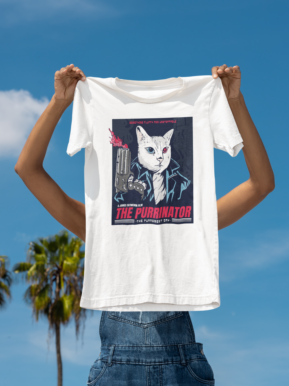 'The Purrinator' t-shirt in white, held up outdoors against a palm tree sky backdrop, funny cat lover tee