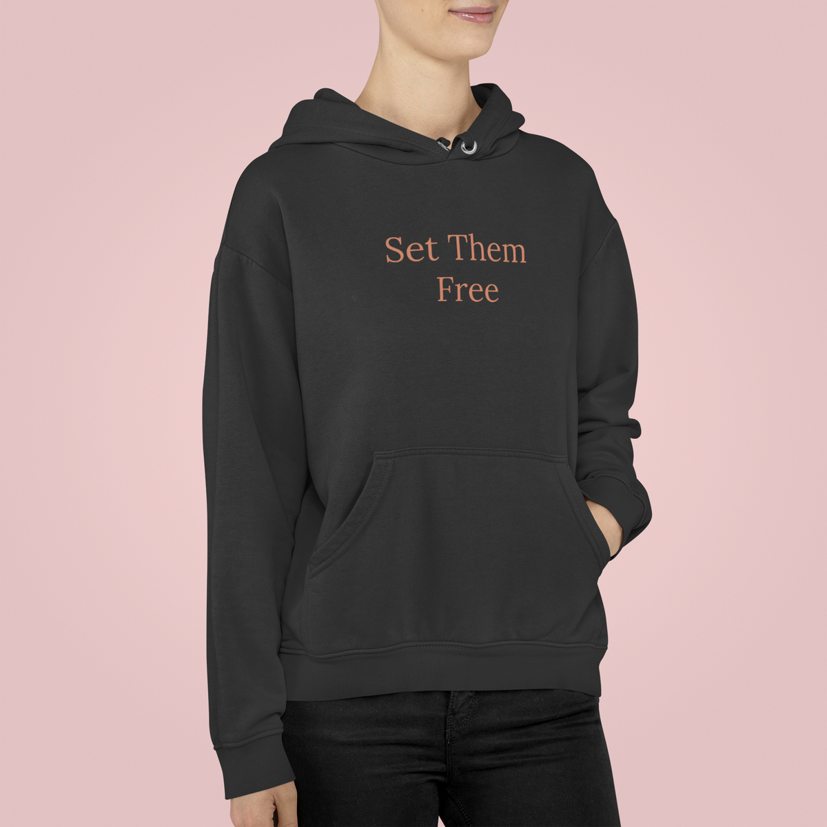 'Set Them Free' hoodie in pink worn by woman on pink backdrop, orange script print, statement gift