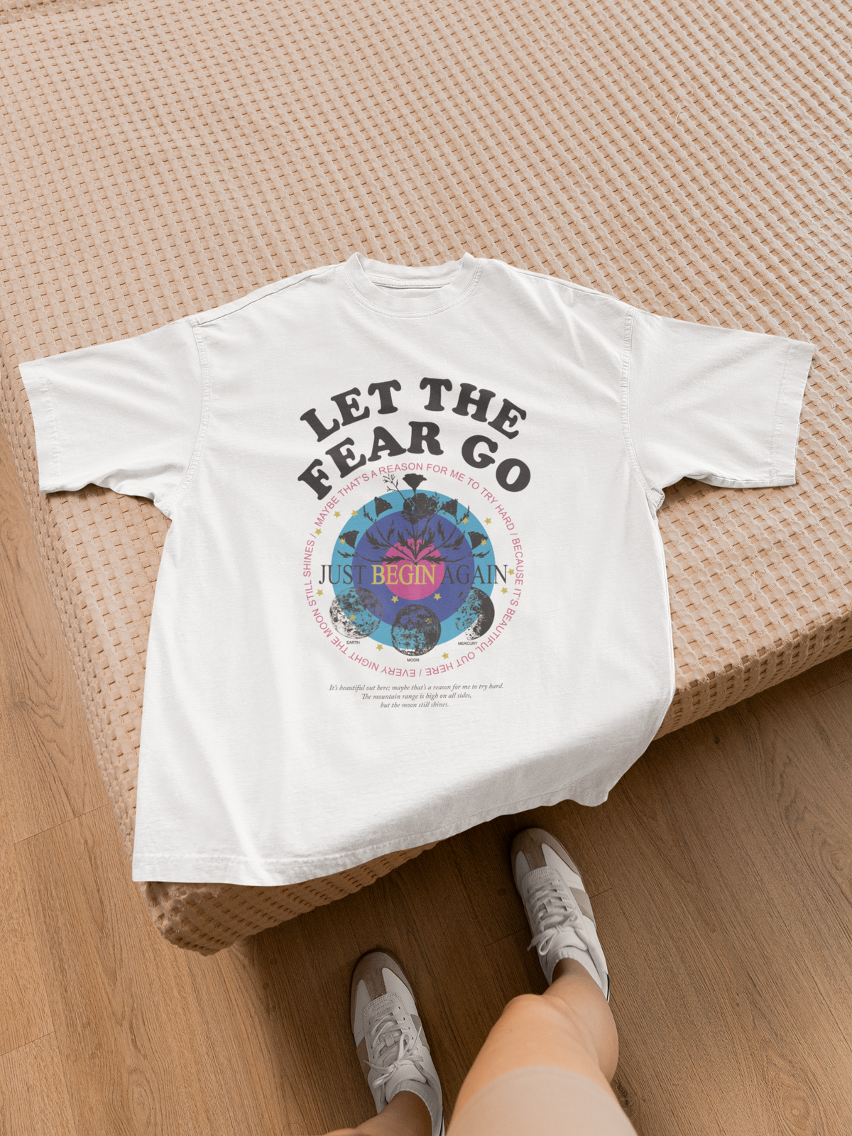 "Let the Fear Go" t-shirt in white, laid out lifestyle shot on bed, inspirational quote graphic tee