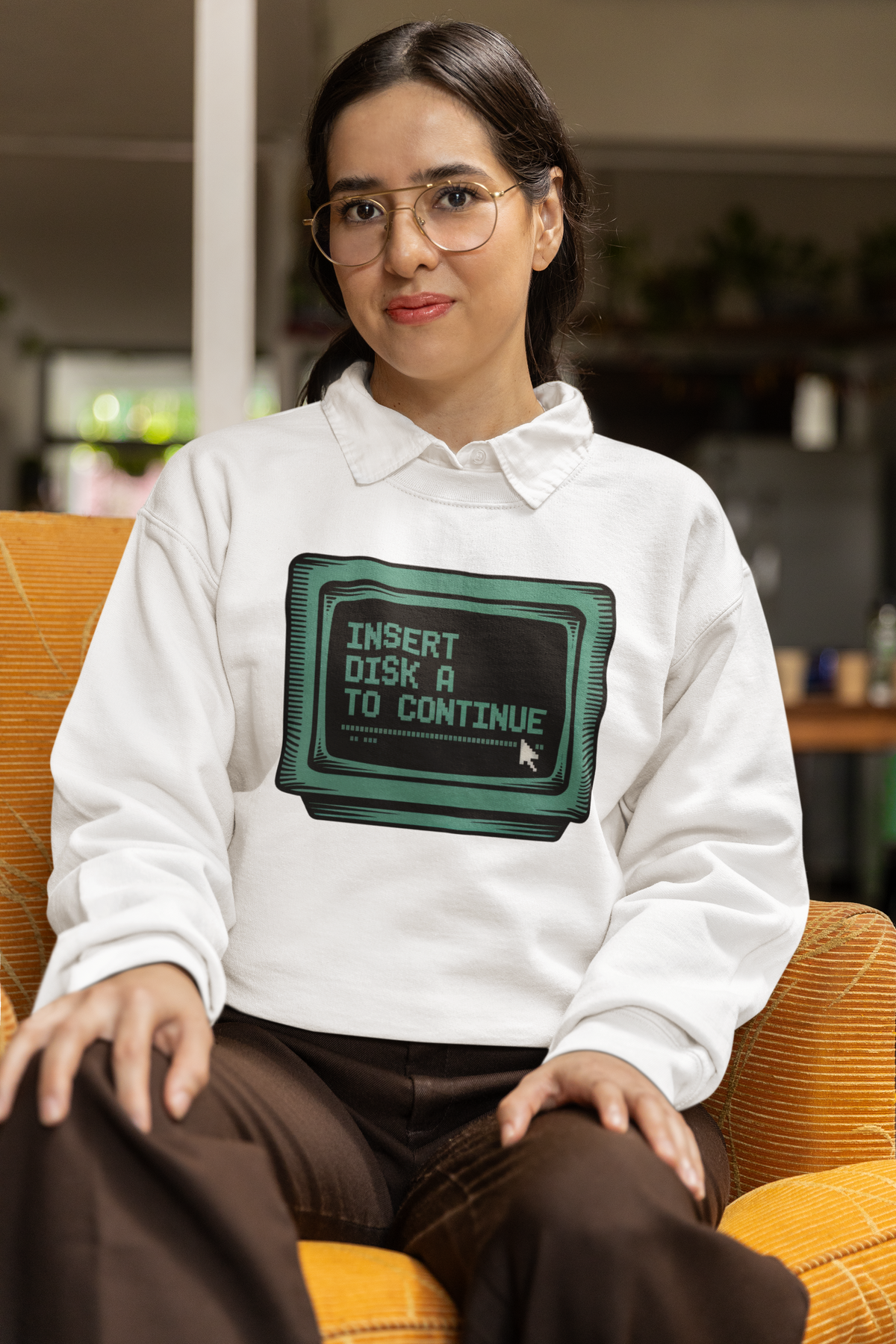 'Insert Disk A to Continue' sweatshirt in white, woman wearing it seated indoors, retro gamer nostalgia gift