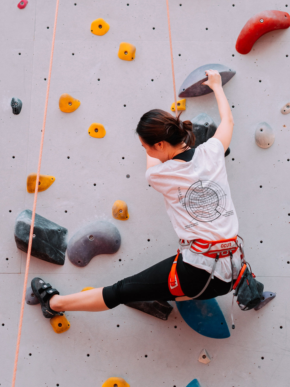 A woman climbing on a rock wall wearing a Spherical Angles T-Shirt in various sizes.
